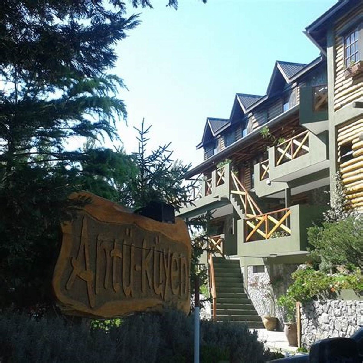 Letrero de madera tallada con el nombre Antü·Küyen, y detrás el edificio de troncos con sus balcones.