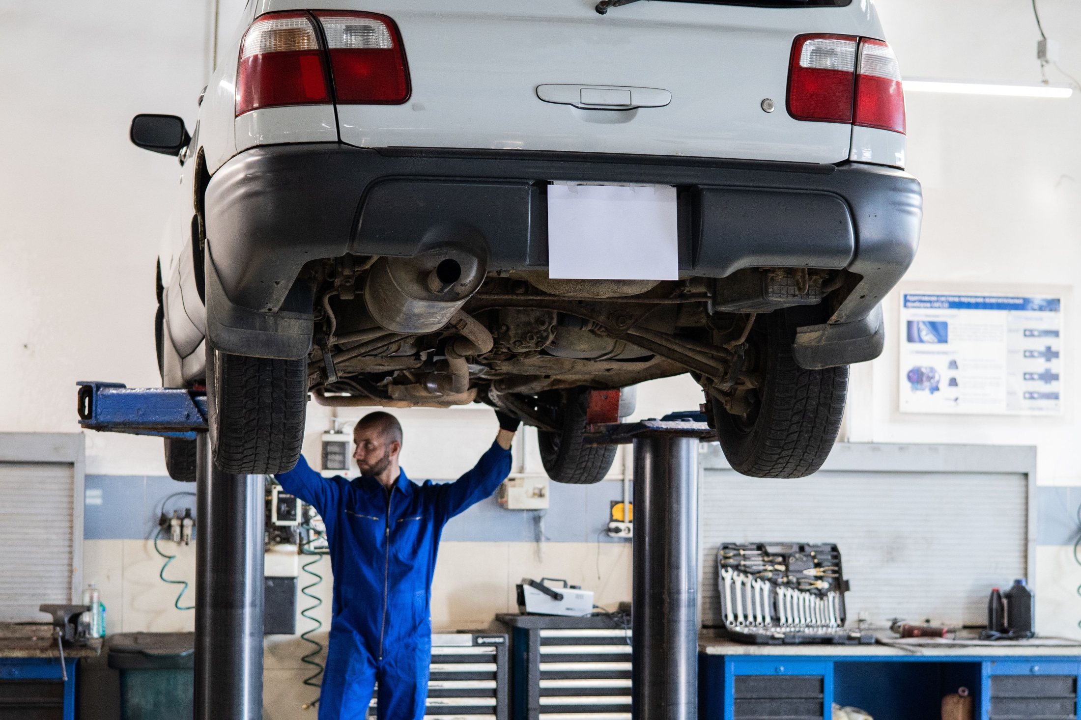 Auto levantado en un alza de dos columnas, el mecánico revisando los bajos y el banco de herramientas al fondo