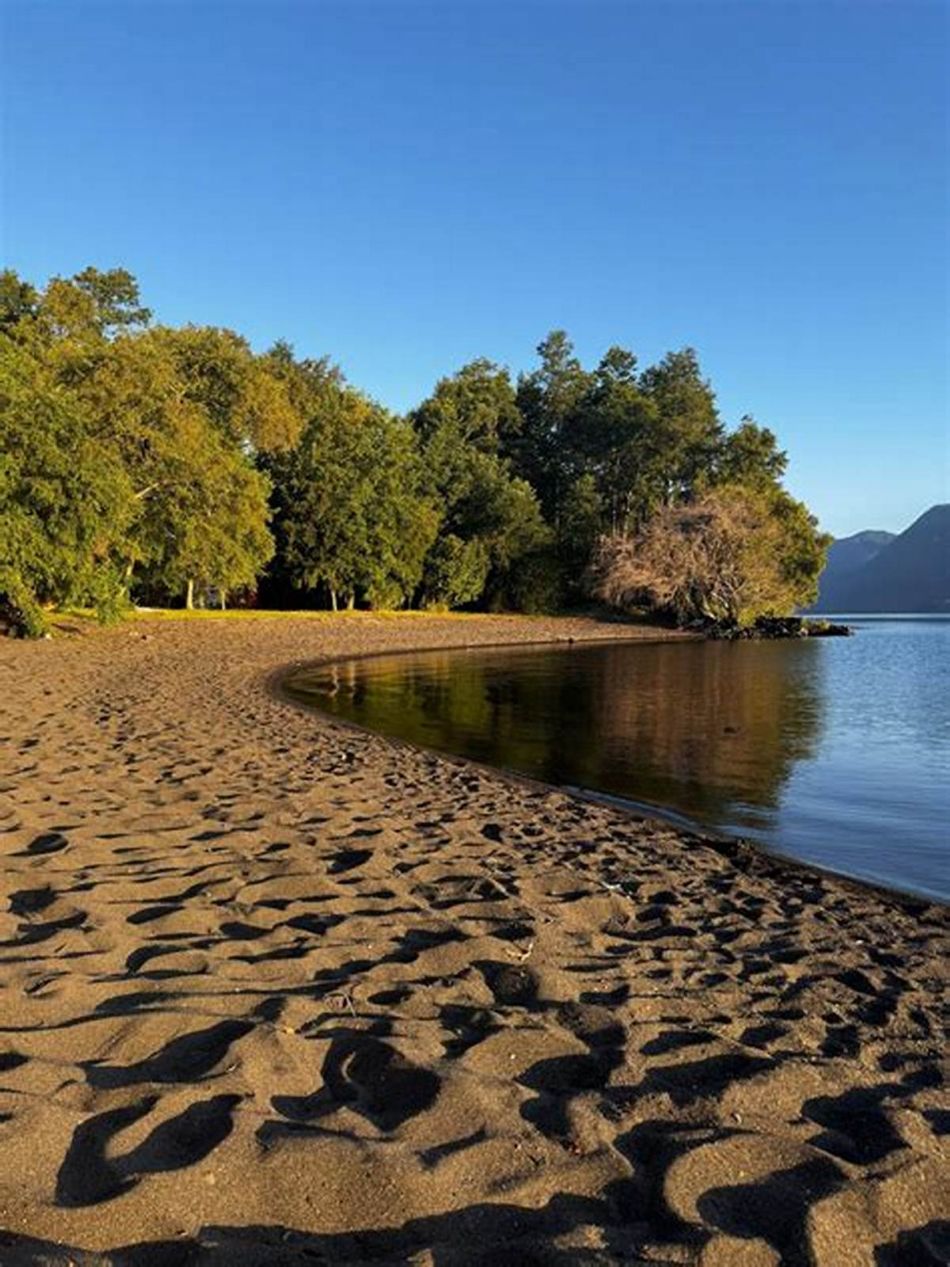 La playa de arena oscura del camping, con el lago y la península al fondo.