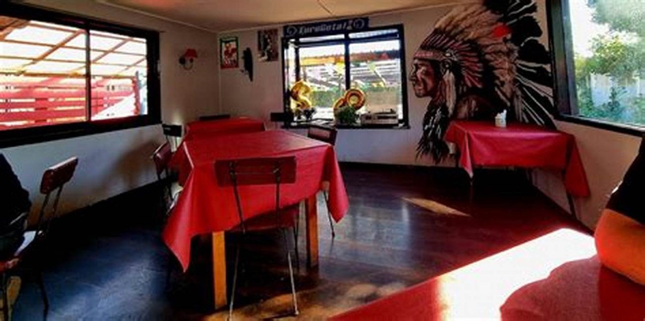 El salón por dentro, con las mesas de manteles rojos y los ventanales al fondo.