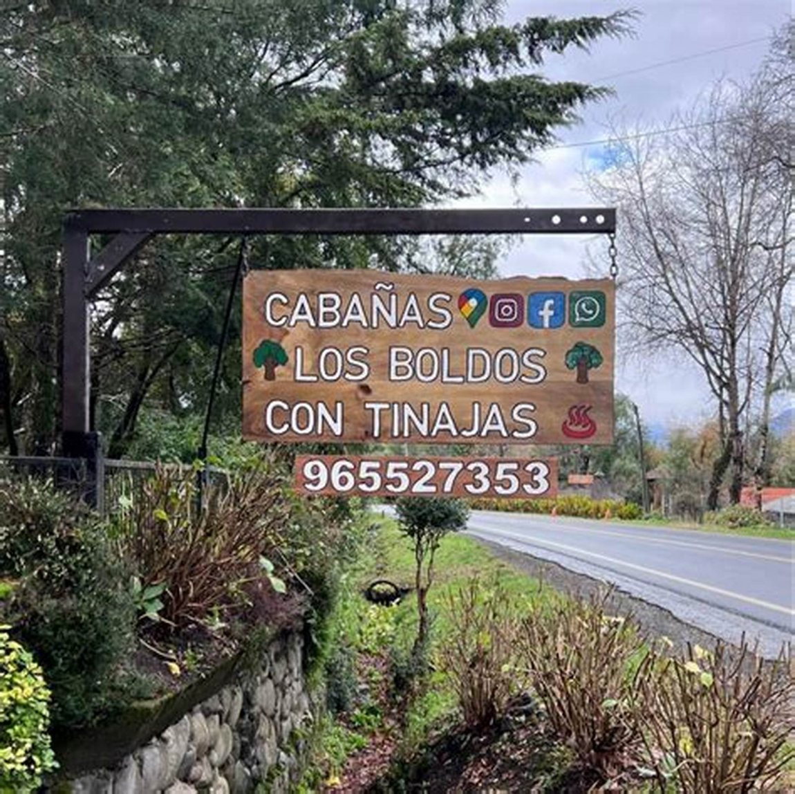 Letrero de madera al borde del camino: Cabañas Los Boldos con tinajas, y el número de teléfono.